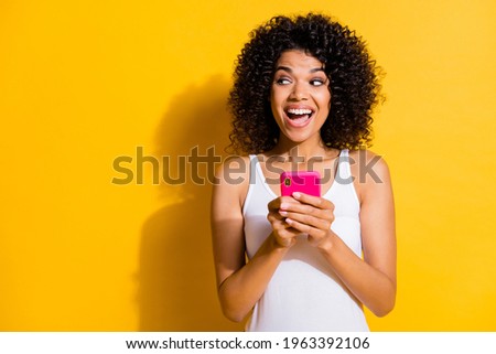 Photo of dark skin brown haired woman look empty space hold phone wear white singlet isolated on yellow color background