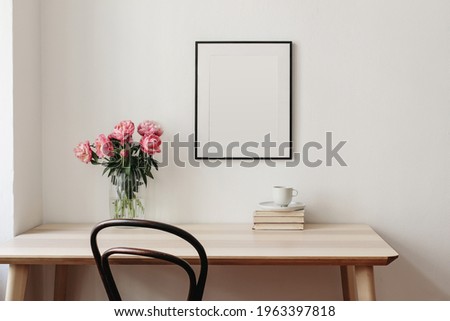 Living room, indoor still life. Empty picture frame mockup on beige wall. Wooden table and old chair. Glass vase with pink peonies bouquet. Elegant working space, home office concept. Interior design.