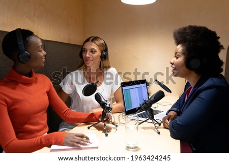 Diverse group of happy female business colleagues wearing headphones, speaking to microphones. recording podcast and business in a modern office.