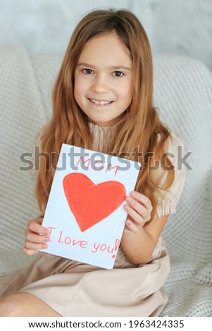 Laughing child girl holding greeting homemade drawing card red heart. Gift for Mothers Day. Happy childhood concept. Inscription Mom, I love you. Attractive daughter congratulate mum, happy birthday