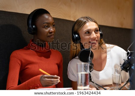 Diverse female business colleagues wearing headphones in discussion speaking to microphones. recording podcast and business in a modern office.