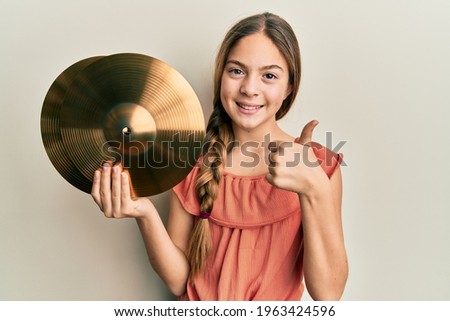 Beautiful brunette little girl holding golden cymbal plates smiling happy and positive, thumb up doing excellent and approval sign 