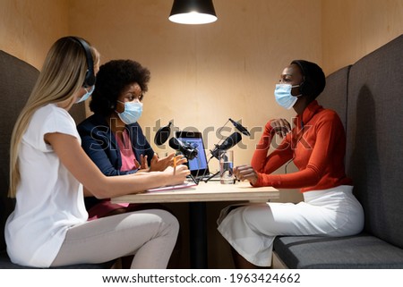 Diverse group of female business colleagues wearing masks in discussion speaking to microphones. recording podcast and business in a modern office during covid 19 coronavirus pandemic.