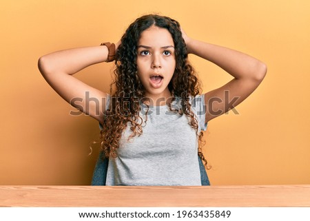 Teenager hispanic girl wearing casual clothes sitting on the table crazy and scared with hands on head, afraid and surprised of shock with open mouth 
