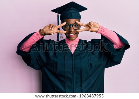 Young african american girl wearing graduation cap and ceremony robe doing peace symbol with fingers over face, smiling cheerful showing victory 