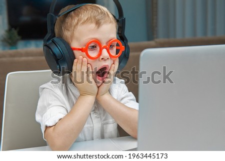 A cute boy in red glasses and wireless headphones is surprised to communicate online using a laptop. Pleasant communication with the use of modern means of communication