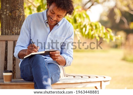 Mature Man With Takeaway Coffee Sitting On Park Bench Under Tree Writing In Notebook Royalty-Free Stock Photo #1963448881