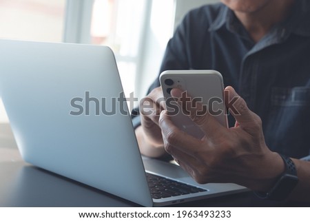 Closeup of business man hand using mobile smart phone and reading message via mobile app, working on laptop computer on office table, online working, business and technology, work from home concept Royalty-Free Stock Photo #1963493233