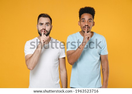 Secret young two friends european african american men 20s in white blue t-shirts say hush be quiet with finger on lips shhh gesture isolated on yellow background studio portrait. Tattoo translate fun