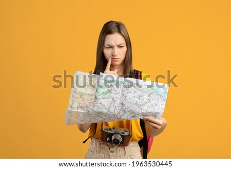 Confused woman traveler with camera looking at city map, got lost, yellow studio background, copy space. Upset young lady backpacker lost in city, checking map. Navigation, trekking during travelling Royalty-Free Stock Photo #1963530445