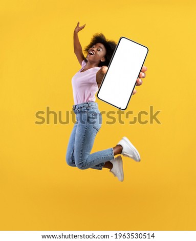 Overjoyed african american young lady showing empty smartphone screen while jumping up over yellow studio background, collage, full size photo. Happy black woman recommending new mobile app Royalty-Free Stock Photo #1963530514