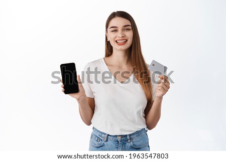 Smiling caucasian woman showing credit card and smartphone screen, recommending mobile application, online shopping store, webpage on phone display, standing over white background