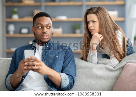 Infidelity Concept. Suspicious Young Woman Watching Her Black Boyfriend Texting On Phone, Jealous Lady Trying To Read Messages While Relaxing In Living Room Together, Suspecting Unfaithfulness Royalty-Free Stock Photo #1963558501