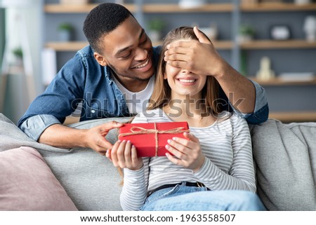 Loving Black Man Covering Eyes And Surprising His White Girlfriend With Romantic Gift At Home, Greeting With Valentine's Day, Birthday Or Anniversary, Mixed Couple Celebrating Holidays Together Royalty-Free Stock Photo #1963558507