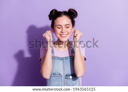 Photo of cute impressed young woman dressed denim overall smiling rising fists closed eyes isolated purple color background
