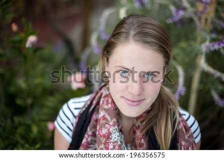 A young blonde woman looks into the camera in front of a green garden background