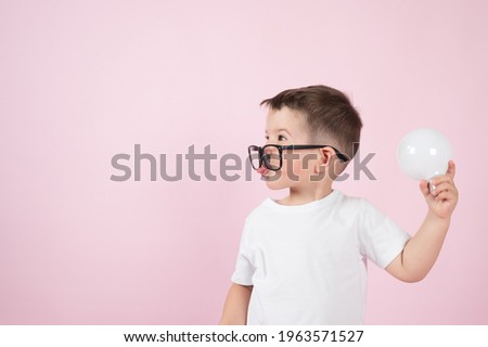 The cute little boy in glasses holds a light bulb and shows his tongue on pink background. The concept of creative thinking. High quality photo