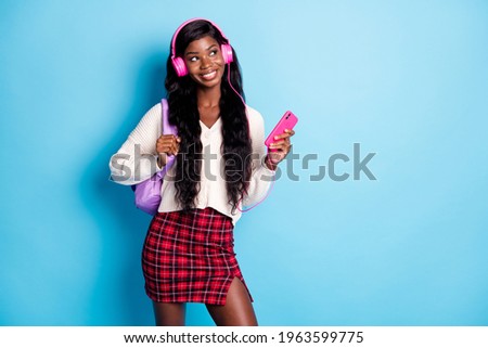 Photo of brunette dark skin dreamy woman look empty space wear headphones casual outfit hold phone bag isolated on blue color background