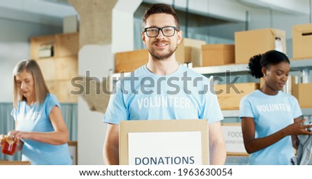 Close up portrait of handsome Caucasian young happy guy volunteer holding in hands donations box, looking at camera and smiling. Charity organization. Women packing donations on background, donating