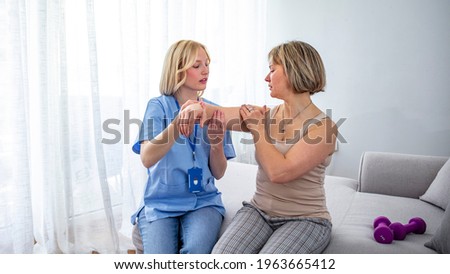 A physiotherapist doing treatment with patient in bright home. Modern rehabilitation physiotherapy. Portrait of female instructor assisting mature woman in lifting dumbbells at home