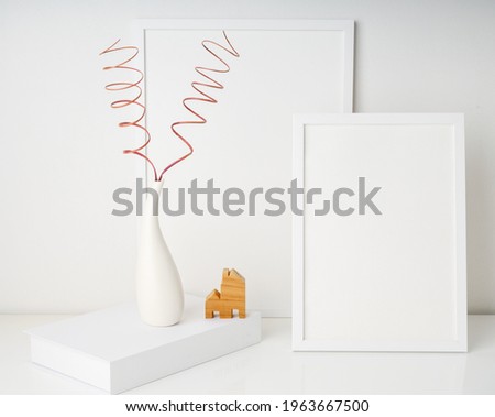 Two mock up white  poster frames decor with dried twings in modern white vase on white book and house model over white table wall background