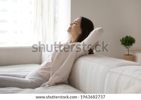 Happy millennial woman relaxing on sofa, breathing cool air from conditioner, enjoying comfortable climate in ventilated living room, meditating with closed eyes. Comfort at home, conditioning concept Royalty-Free Stock Photo #1963682737