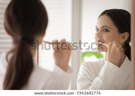 Young woman in bathrobe brushing teeth with wooden bamboo toothbrush. Teen girl choosing eco friendly tools for morning bath routine, oral mouth hygiene, protection from caries. Mirror reflection Royalty-Free Stock Photo #1963682776