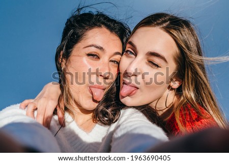 Close-up of a couple of young girls, one blonde and one brunette sticking out their tongues while taking a selfie.
