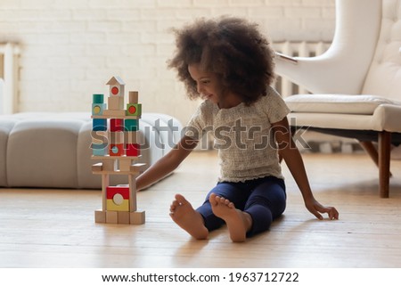 Happy cute African American girl sitting on heat floor, building tower from wood bricks and blocks. Kid playing active games at home, improving construction and architecture skills. Playtime concept Royalty-Free Stock Photo #1963712722