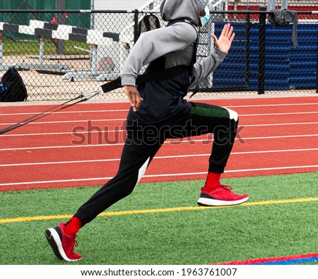 A male high school runner wearing a protective face mask is dragging a sled with weight across a green turf field during strength and agility practice.