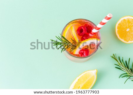 Cool summer drink with lemon, raspberry and rosemary in glass with striped eco straw on green background. Bright cold fruit cocktail, top view with copy space.