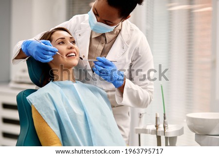 African American dentist examining teeth of her female patient during appointment at dental clinic. Focus is on young woman. Royalty-Free Stock Photo #1963795717