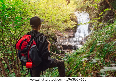 young girl standing with backpack observing a forest landscape with waterfall. woman in front of a waterfall enjoying the nature. Active turism. Royalty-Free Stock Photo #1963807663
