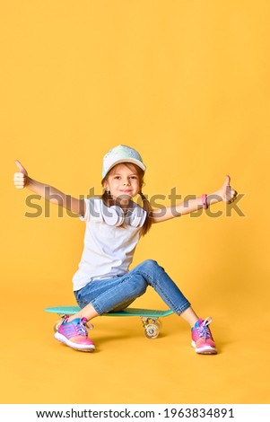 Stylish funny girl wearing white t-shirt, blue jeans and sneakers, sitting on skateboard over yellow background and raises hands and thumbs up emotionally.