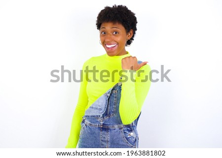 Impressed African American female with curly bushy wears jeans overalls over white wall point back empty space