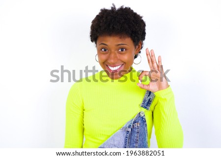 African American female with curly bushy wears jeans overalls over white wall hold hand arm okey symbol toothy approve advising novelty news