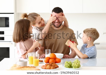 Surpise for daddy. Cute smiling children siblings giving father wrapped gift box and postcard. Young mom congratulating husband with kids while having breakfast in kitchen. Family holiday concept Royalty-Free Stock Photo #1963960552