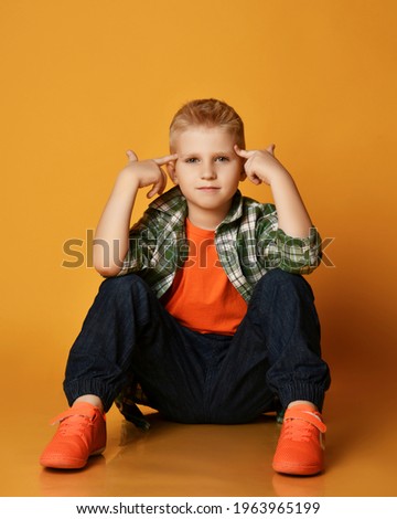 Clever schoolboy teenager in checkered shirt, orange t-shirt and blue jeans sits on floor holding fingers at head, gesturing think it over, keep in mind gesture over yellow background