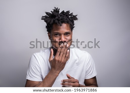An Afro American young fit athletic man, in the white t-shirt, covering his mouth with his hand, laughing hard, bursting into laughter, isolated on the grey background