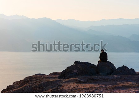 A man sits on a high mountain cliff and overlooks the horizon. A concept for hope, peace, power, soul searching. Desert mountains against the backdrop of the Red Sea. Shlomo mountain, Eilat Israel Royalty-Free Stock Photo #1963980751