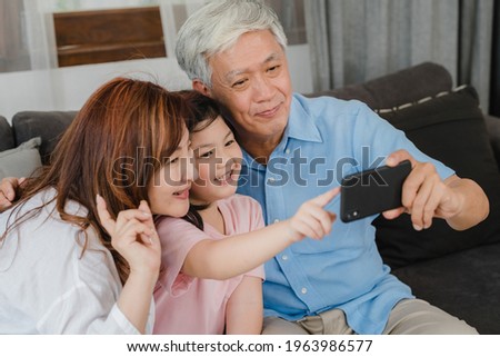 Asian grandparents selfie with granddaughter at home. Senior Chinese, grandpa and grandma happy spend family time relax using mobile phone with young girl kid lying on sofa in living room concept.