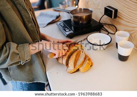 Woman cooking on a high kitchen table, she's cutting bread on a board while rise is boiling on a stove. Cropped, no head. Side view. Royalty-Free Stock Photo #1963988683