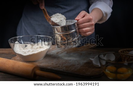 Women's hands, flour and dough. A woman, in an apron prepares dough for homemade baking, a rustic home cozy atmosphere, a dark background with unusual lighting.