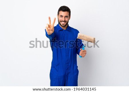 Painter man holding a paint roller isolated on white background smiling and showing victory sign