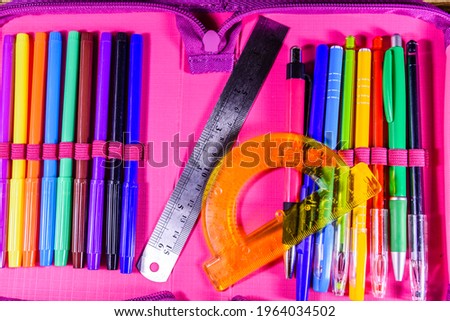 Different school stationeries (pens, pencils, felt tip pens, ruler and protractor) in pink pencil box. Top view