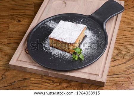 Freshly baked apple pie with apples, mint leat and powdered sugar. Served on a black plate over wooden background. Sweet dessert, one piece.