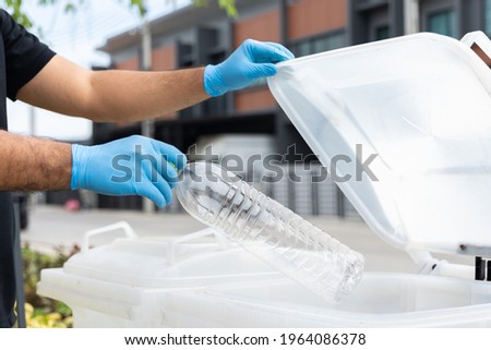 A young man wearing rubber gloves holds plastic bottles in the trash in front of his house.