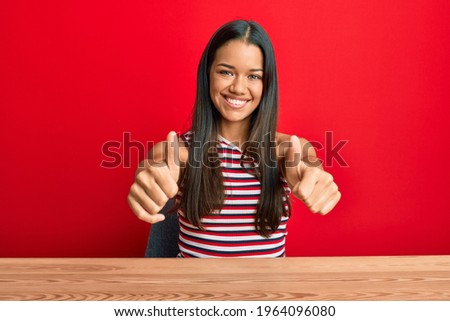 Beautiful hispanic woman wearing casual clothes sitting on the table approving doing positive gesture with hand, thumbs up smiling and happy for success. winner gesture. 