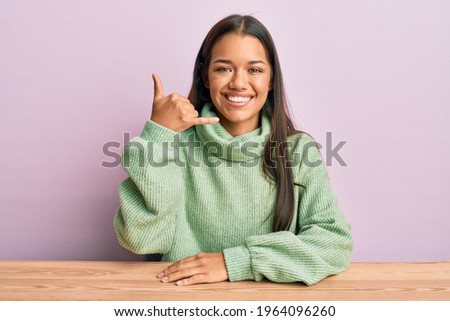 Beautiful hispanic woman wearing casual clothes sitting on the table smiling doing phone gesture with hand and fingers like talking on the telephone. communicating concepts. 