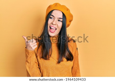 Young brunette woman wearing french look with beret pointing thumb up to the side smiling happy with open mouth 